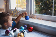Happy little child boy holding blue Christmas ball near window indoor with warm garland lights on blurred background.