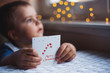 Child boy holding Christmas greeting card with warm garland bokeh on background near window in daylight.