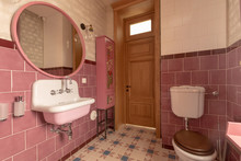 Interior Of Clean Light Restroom With Retro Sink Beside Big Round Mirror And Decorated Cabinet On Tall Wall With White And Pink Tile Next To Toilet Bowl With Brown Lid In Apartment