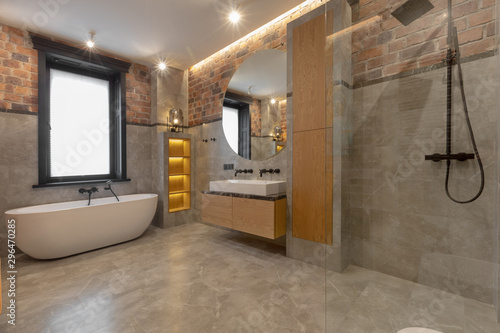 Loft interior of spacious wet room with bathtub near window and open walk in shower beside cabinet and sink with mirror against decorated built in shelves on gray marble