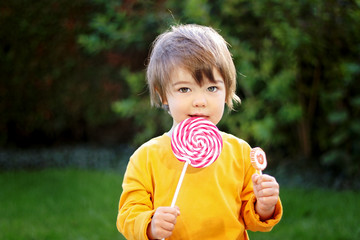 Wall Mural - Portrait of little boy with funny hair eating big red lollipop outdoors on green grass looking at camera. Sweet tooth. Carefree childhood. Summer lifestyle