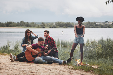 Wall Mural - Fire is burning, music is playing. Group of people have picnic on the beach. Friends have fun at weekend time