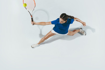 Wall Mural - Young woman in blue shirt playing tennis. She hits the ball with a racket. Indoor studio shot isolated on white. Youth, flexibility, power and energy. Negative space. Top view.