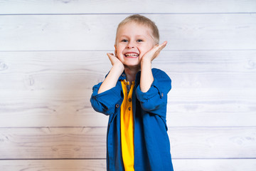 Portrait of a beautiful kid boy in yellow T-shirt and denim jacket, shirt. Boy standing on a white wooden background. 5 years old boy. Hands near face.