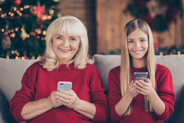 Sticker - Close-up portrait of nice attractive charming cheerful cheery granny pre-teen long-haired grandchild sitting on divan using cell sending sms congrats at decorated industrial loft style interior house