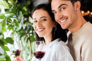 Wall Mural - selective focus of handsome man holding glass with red wine near positive girlfriend