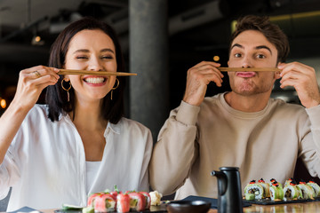 Wall Mural - handsome man holding chopsticks near face while sitting with cheerful girl in sushi bar