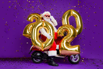 Wall Mural - Full body photo of fat santa man sitting on bike holding big air newyear 2020 numbers balloons confetti falling wear sun specs x-mas costume isolated purple background