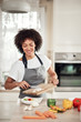 © nenadaksic - Smiling mixed race woman in apron putting chopped mushrooms in bowl with vegetables. Kitchen interior.