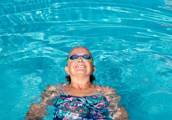 An elderly woman with swimming pool goggles in the transparent  water of the swimming pool. Smiles and happiness.. Healthy activity for pensioner.