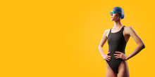 Young Female Swimmer Portrait Of Half Body In Black Swimsuit With Goggle And Swimming Hat. Akimbo Posing In Studio. Isolated On Yellow Background