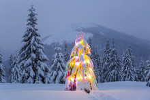 The Christmas Tree Decorated With Colourful Garlands Stands On The Snowy Lawn. Night Winter Mountain Landscapes.