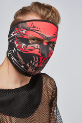 Close-up portrait of a blond man, posing on a grey background. He is wearing black mesh tank top. His face is hidden by  black full face mask with red skull print. His the hair is combed back. 