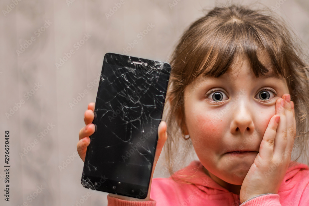 Portrait of a scared little girl with a broken mobile phone. A sad child broke the screen of a mobile phone. Cracked display in hand kids. The frightened child accidentally ruined the smartphone - obrazy, fototapety, plakaty 