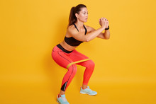 Studio Shot Of Sporty Woman Squatting, Doing Sit Ups With Resistance Band. Photo Of Caucasian Woman In Fashionable Sportswear Isolated Over Yellow Background. Strength And Motivation Concept.