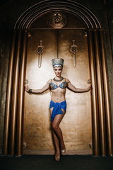 Wall Mural - Beautiful thin Egyptian woman with pronounced cheekbones in a huge gold headdress without hair in a blue swimsuit on the background in the Studio