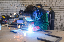 A Strong Man Is A Welder In A Welding Mask And Welders Leathers, A Metal Product Is Welded With A Welding Machine In The Garage, Blue Sparks Fly To The Sides