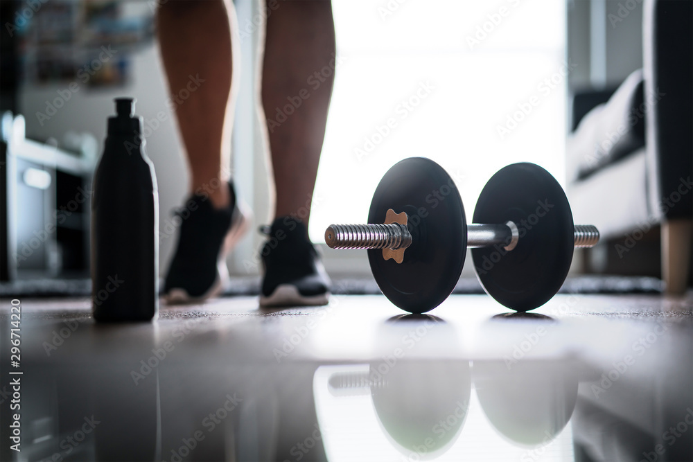 Fitness, home workout and weight training concept. Man starting to exercise with dumbbell. Water or protein shake bottle on floor. - obrazy, fototapety, plakaty 