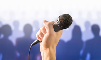 Public speaking and giving speech concept. Hand holding microphone in front of a silhouette audience and crowd of people. Singing to mic in karaoke or talent show concept.