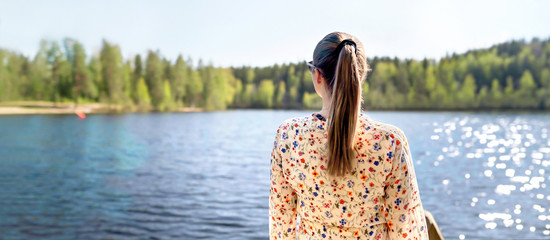 Finnish woman standing and looking at a lake in Finland. Happy person relaxing and enjoying sunny summer vacation or weekend in nature. Back view of carefree lady in dress. Wide banner.