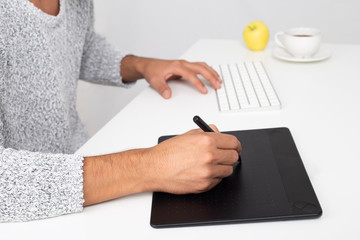 young graphic designer working in his computer in a desktop. He is using a graphic tablet. He is wearing a gray sweater. On the desk there is a cup of coffee and an apple