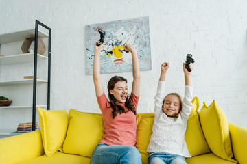 Wall Mural - KYIV, UKRAINE - SEPTEMBER 4, 2019: cheerful babysitter and happy kid celebrating triumph while holding joysticks in living room