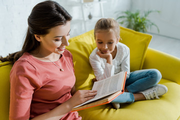 Wall Mural - selective focus of happy kid sitting on sofa with happy babysitter reading book