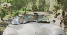 Happy Smiling Diverse Newly Wed Couple Driving On A Sunny Country Road, Bride And Groom Driving Together In A White Vintage Convertible, Bride Holding Flower Bouquet