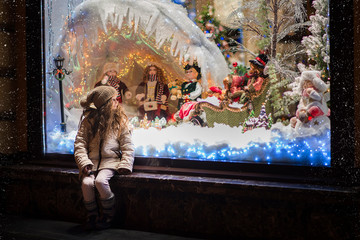  Little girl stands on the streets at the window on Christmas Eve