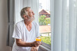 © wirojsid - Senior elderly man standing and looking out of window in bedroom after waking up in morning with juice