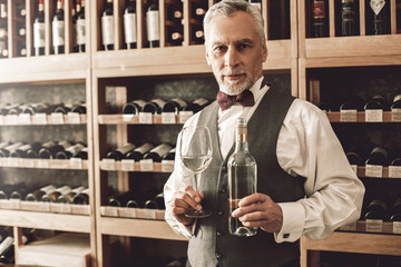 Wall Mural - Sommelier Concept. Senior man standing with bottle and glass of white wine smiling joyful