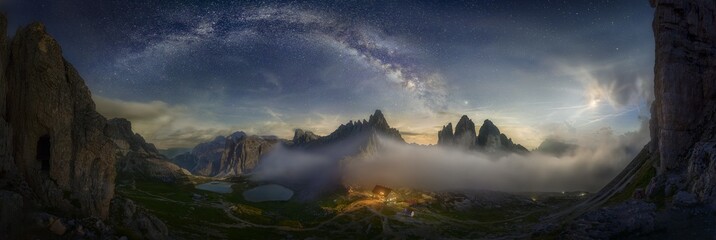 Wall Mural - Beautiful panoramic shot of a mountain valley with a small illuminated house under the starry sky