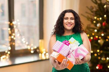 Wall Mural - holidays, presents and people concept - happy woman in green dress holding gift boxes over christmas tree lights on home background