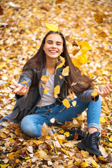 Young girl in blue jeans and gray coat sits on autumn leaves in a park