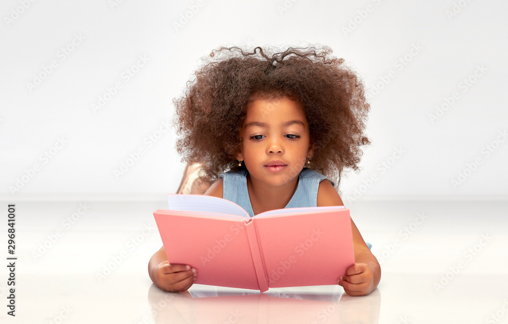 childhood, school and education concept - little african american girl reading book over grey background - obrazy, fototapety, plakaty 