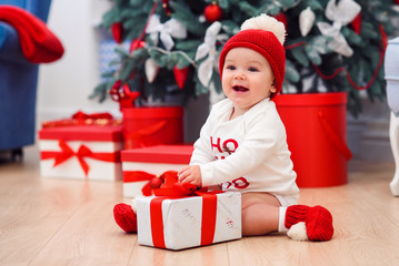 Charming toddler boy holds white christmas gift box with red ribbon. Funny cute baby weared in festive clothes in Christmas decorated room. Concept of Christmas and New Year holidays.