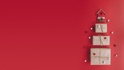 Photo of gift boxes in kraft paper in the shape of a fir tree with wooden house on the top. Near with box xmas bells. Red background. Flat lay Christmas composition. Nativity mood. Copy space, 16:9.