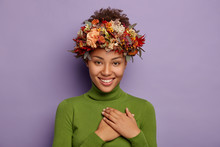 Candid Shot Of Attractive Smiling Woman Feels Thankful, Promises Be Loyal, Appreciates Pleasant Gift, Touches Chest Gratefully, Wears Autumn Wreath, Values Somebodys Offer, Poses Indoor In Casual Wear