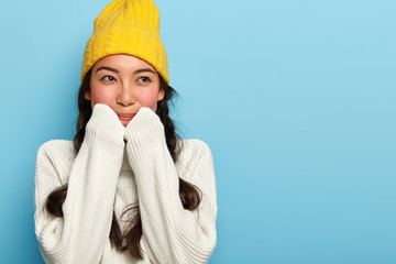 Indoor shot of lovely brunette woman wears yellow hat and white sweater, ready for winter stroll with boyfriend, has dreamy expression, looks away as recalls pleasant moment, stands against blue wall