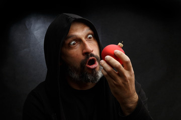 Wall Mural - Portrait of a bald man with a beard in a black hood witha red christmas ball on a dirty gray background. Young Santa concept.