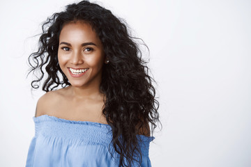 Wall Mural - Beauty, wellbeing and women concept. Charming curly-haired african american girl in blue blouse smiling joyfully, gaze with happy tender expression camera, standing white background