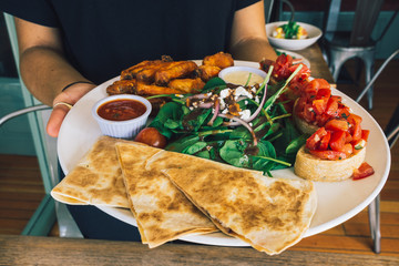 Women holding a platter with bruschetta, buffalo wings and quesadilla over a wooden table.