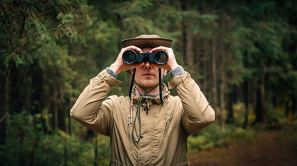 A man in a hat and uniform green and beige holds binoculars and looks into the distance, Ranger watching the territory, the protection of the reserve