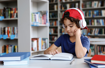 Wall Mural - Cute little boy with headphones reading books at table in library