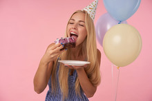 Portrait Of Young Delighted Blonde Female In Holiday Outfit Holding Big Piece Of Delicious Cake And Going To Eat It, Celebrating Birthday With Multicolored Air Balloons, Posing Over Pink Background