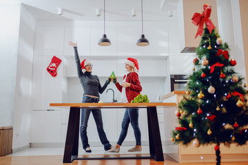 Wall Mural - Overjoyed caucasian senior woman and her daughter toasting with beer in kitchen on christmas eve. Both having santa hats on head. On kitchen counter are vegetables.