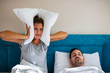  Woman suffers from her male partner snoring in bed. Noise concept. Real people. Copy space. Young irritated woman lying in bed with snoring husband at home