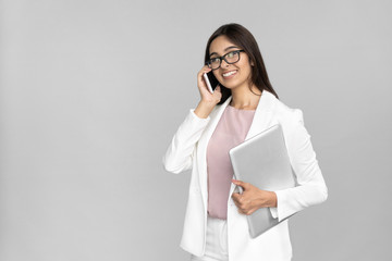 Cheerful and happy young adult indian business lady in white suit talk on smartphone holding modern laptop in hand. Smiling woman stand isolated on grey background, copy space, corporate tech concept