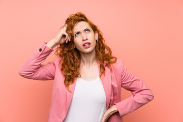 Wall Mural - Redhead woman in suit over isolated pink wall having doubts and with confuse face expression