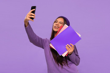 Pretty african-american teenager student in a hat and sweater holding folders and making selfie with a smartphone standing isolated over purple background.